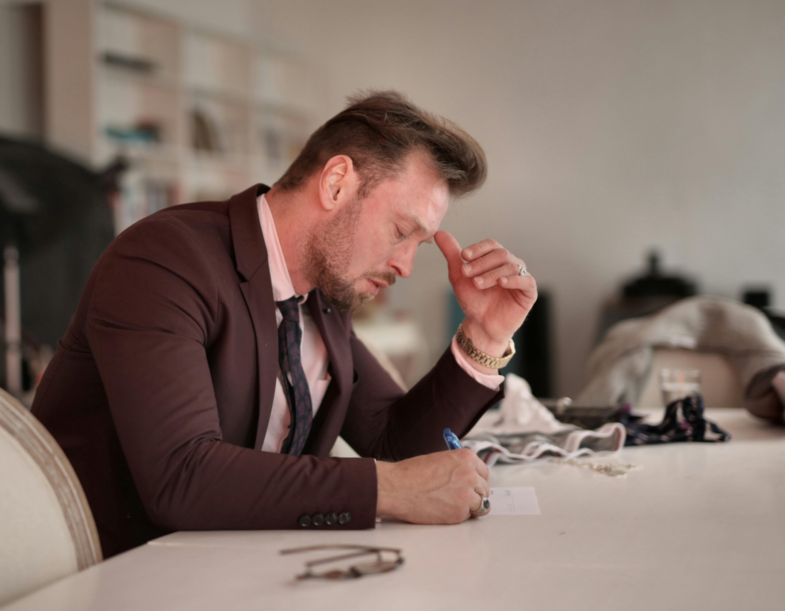 Side view of brutal male entrepreneur in formal suit sitting at table while contemplating and writing thoughts on paper